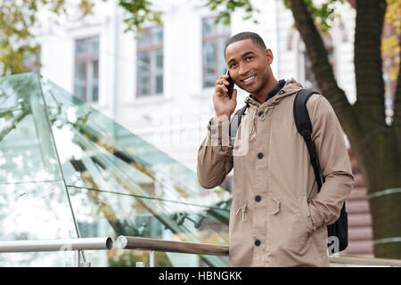 Picture of smiling young african man talking on his phone on the street. Looking aside. Stock Photo