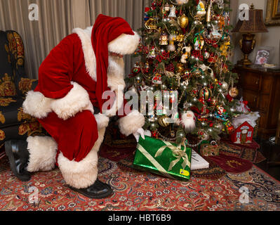 Santa Claus kneeling placing gift under decorated Christmas tree Stock Photo