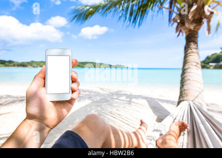 Using smartphone on a tropical paradise beach sitting in hammock, mobile phone and technology during sea holidays Stock Photo