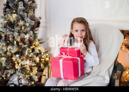Happy girl opening Christmas gifts by a decorated fireplace in cozy light living room on Xmas eve Stock Photo