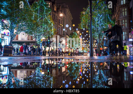 LONDON - NOVEMBER 16, 2016: Holiday lights twinkle in a puddle reflection as pedestrians crowd the sidewalks of Oxford Street. Stock Photo