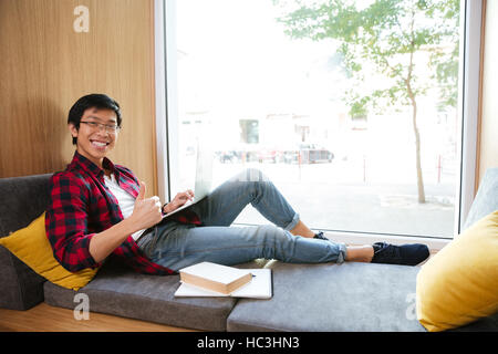 Image of happy asian student using laptop in university library sitting on sofa near book and notebook. Stock Photo
