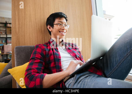 Photo of joyful asian student wearing eyeglasses and dressed in shirt in a cage using laptop in university library sitting on sofa. Looking at laptop. Stock Photo