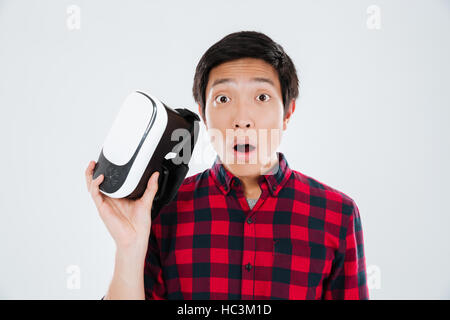 Picture of young shocked man dressed in casual shirt in a cage and holding virtual reality device. Isolated over white background. Stock Photo