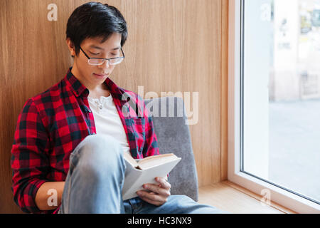 Photo of cheerful asian student reading a book in university library sitting on sofa. Stock Photo