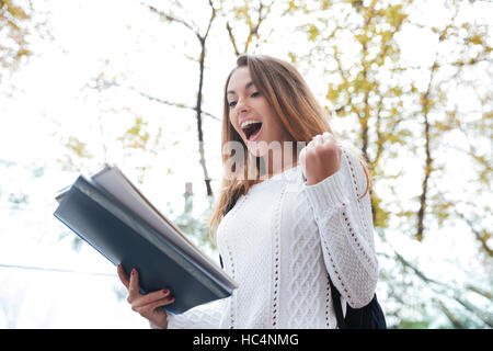 Cheerful excited young woman with notebook shouting and celebrating success outdoors Stock Photo