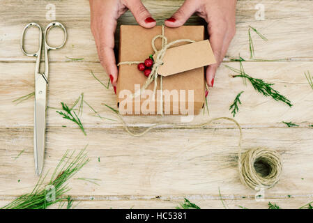 closeup of a young caucasian woman with a freshly arranged christmas gift in her hands, on a rustic wooden with some twigs, a pair of scissors and a r Stock Photo
