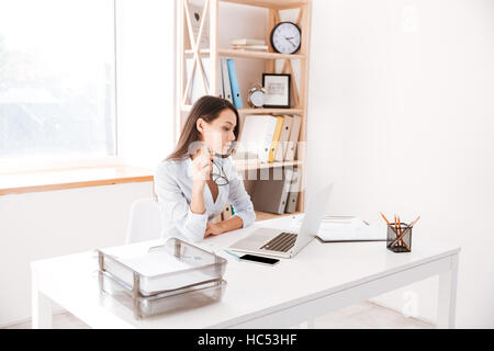 Concentrated businesswoman dressed in white shirt sitting in her office and looking at laptop while holding eyeglasses. Stock Photo