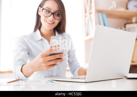 Cheerful businesswoman dressed in white shirt sitting in her office and looking at phone. Focus on cellphone. Stock Photo