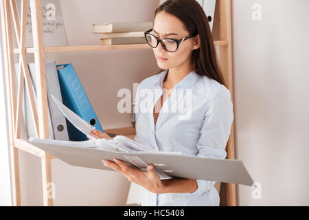Young concentrated businesswoman holding folder near book shelf while standing in office Stock Photo