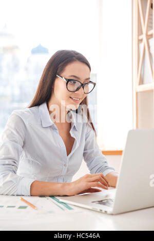 Photo of businesswoman dressed in white shirt and wearing glasses sitting in her office and using laptop. Looking at laptop. Stock Photo