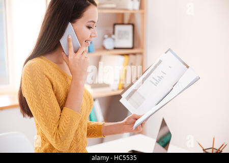 Photo of beautiful businesswoman standing and looking at documents while talking by phone Stock Photo
