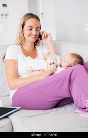 Mother talking on mobile phone while feeding her baby with milk bottle Stock Photo