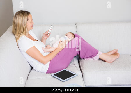 Mother using mobile phone while feeding her baby with milk bottle Stock Photo