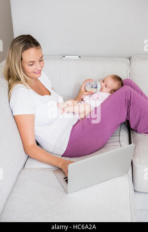 Mother using laptop while feeding her baby with milk bottle Stock Photo