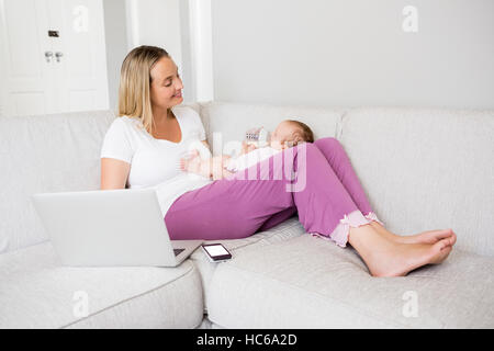 Mother using laptop while feeding her baby with milk bottle Stock Photo