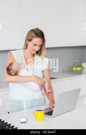 Mother using laptop while carrying baby in kitchen Stock Photo