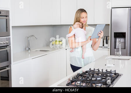 Mother using digital tablet while carrying her baby in kitchen Stock Photo