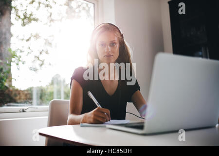Portrait of young woman sitting in kitchen with headphones writing note. Female studying at home with laptop. Stock Photo