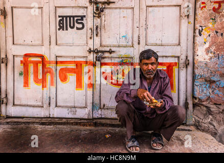 A middle aged Indian man stoops in front of a white door with red hindi symbols looking directly ahead with food in his hands. Stock Photo