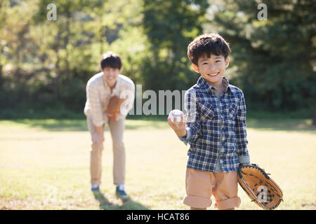 Smiling five-year-old boy with glove and baseball standing on grassland with his father behind him Stock Photo