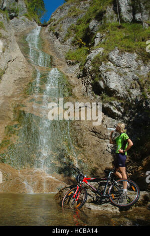 22 year-old woman, on mountain bike wearing cycle clothing, standing by a waterfall, Kalkalpen National Park, Upper Austria Stock Photo