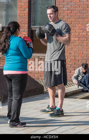 A man wearing boxing gloves is demonstrating how to box to a woman who is also wearing gloves. A mature woman is sitting on the ground behind smoking. Stock Photo