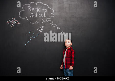 Image of young cute schoolboy standing over background of black chalkboard with school drawings. Look at drawings. Stock Photo