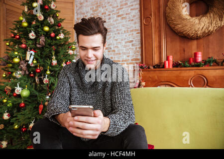 Man in sweater sitting on sofa with phone near the fir-tree Stock Photo