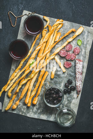 Wine and appetizers set. Italian Grissini bread sticks, dry cured pork meat sausage, black olives in jar and red wine in glasses over dark stone backg Stock Photo