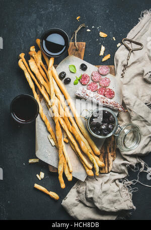 Wine and appetizers set. Italian Grissini bread sticks, dry cured pork meat sausage, black olives on rustic wooden serving board and red wine in glass Stock Photo