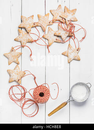 Christmas homemade gingerbread star shaped cookies with sugar powder in sieve and red decoration rope over white wooden background, top view, copy spa Stock Photo