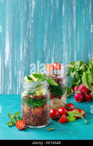 Salads with quinoa, couscous, spinach, radish, tomatoes and zucchini in glass mason jars, standing with fresh vegetables over bright turquoise wooden Stock Photo