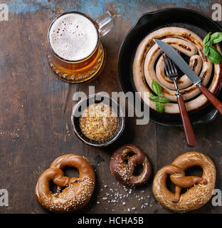 Glass of lager beer with fried sausage in pan with cutlery and traditional salted pretzels and bowl of mustard over old dark wooden background. Top vi Stock Photo