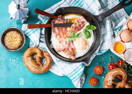 Breakfast with fried eggs and bacon in cast-iron pan, broken egg, tomatoes, mustard, pretzels and basil on white kitchen towel over turquoise wooden b Stock Photo