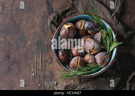 Roasted chestnuts in the ashes with rosemary in turquoise ceramic bowl on sackcloth over old dark wooden background. Top view with space for text. Stock Photo