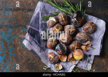 Roasted chestnuts in the ashes with rosemary on paper over old dark texture wooden background. Top view with space for text. Stock Photo