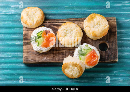 Sliced traditional english cheese scones with smoked salmon, creme cheese and fresh cucumber served with whole scones on wooden chopping board over tu Stock Photo