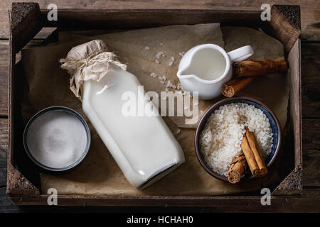 Ingredients for making rice pudding. Bowl of white uncooked rice, sugar, cinnamon sticks, bottle of milk and jug of cream in wood box over old wooden Stock Photo