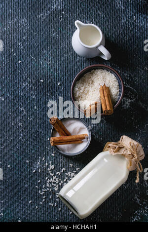 Ingredients for making rice pudding. Bowl of white uncooked rice, sugar, cinnamon sticks, bottle of milk and jug of cream over black texture backgroun Stock Photo