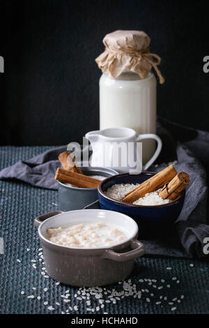 Ingredients for making rice pudding. Bowl of white uncooked rice, sugar, cinnamon sticks, bottle of milk and jug of cream and pot of cooking pudding o Stock Photo