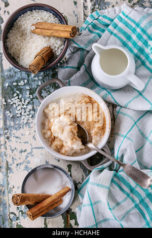 Ingredients for making rice pudding. Uncooked rice, sugar, cinnamon sticks, milk, jug of cream and pot of cooking pudding on kitchen towel over white Stock Photo
