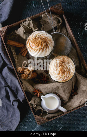 Pumpkin spicy latte  with whipped cream and cinnamon in two glasses standing in wooden box with textile, jug of cream and spices other dark background Stock Photo