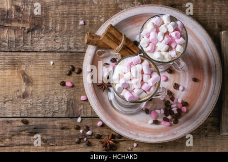 Two glasses of cafe latte with pink marshmallow standing on ceramic plate with spices, coffee beans and pink sugar over wooden texture background. Top Stock Photo