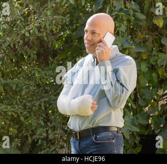 Young man with an arm and elbow in a white plaster / fiberglass standing in a garden, happily talking on his phone Stock Photo
