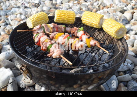 Mini compact barbecue on a stone beach cooking chicken skewers with sweetcorn Stock Photo