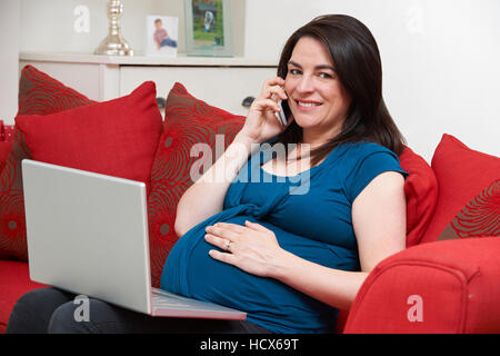 Pregnant Woman Sitting On Sofa Using Laptop And Mobile Phone Stock Photo