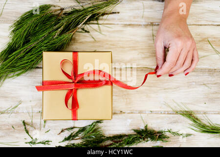 high-angle shot of a young caucasian woman with her fingernails painted red tying a red ribbon around a gift wrapped in a golden paper, on a rustic wo Stock Photo