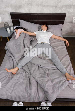Young handsome man sleeping in free fall position with his girlfriend occupied the whole bed, wearing pajamas, near bedside table with candles Stock Photo