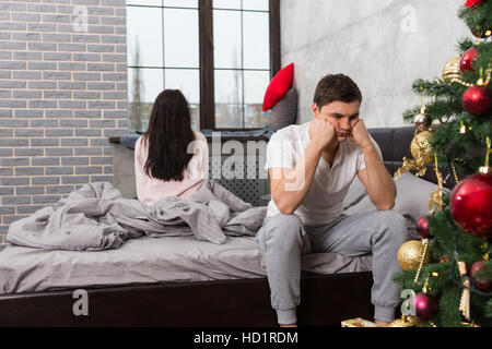 Young man is leaning on his hands while sitting sadly on bed, girlfriend is sitting on bed and looking out the window in the background Stock Photo